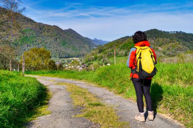Sarı bir sırt çantasıyla toprak yolda yürüyen bir kadın. Yol dağlarla çevrili ve gökyüzü temiz. Kadın manzaranın ve bölgenin huzurunun tadını çıkarıyor.