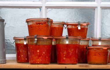 Kırmızı sosla dolu cam kavanozlar. Kavanozlar üst üste istiflenmiş, bolluk ve çeşitlilik hissi yaratıyorlar. Sahne bir sıcaklık ve rahatlık hissi veriyor.