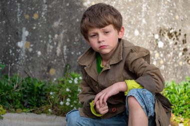 Genç bir çocuk elleri dizlerinin üzerinde yerde oturuyor. O, kahverengi bir ceket ve yeşil bir gömlek giyiyor. Çocuk üzgün ya da üzgün görünüyor.