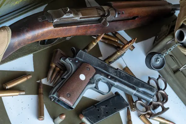 Masada silah ve bir sürü mermi var. Silah bir tüfek ve mermiler bir yığın içinde. Olay yeri gergin ve ciddi. Silah ve mühimmatı gösteriyor.