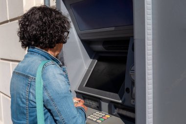 Kadın ATM makinesi kullanıyor. O, mavi bir ceket giyiyor. ATM 'nin siyah ekranı var.