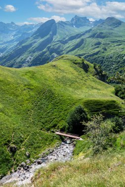 Yeşil bir yamacı ve nehir üzerinde köprüsü olan bir dağ sırası. Köprü ahşaptır ve dağ sırasının ortasında yer alır.