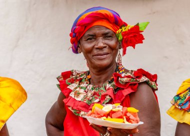 PALENQUERA, CARTAGENA, COLOMBIA, 20 Şubat 2024: Eski Cartagena kasabasındaki neşeli taze meyve sokak satıcısı namı diğer Palenquera. Geleneksel giyinmiş mutlu Afro-Kolombiyalı kadın, Kolombiya kültürü.