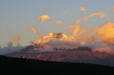 Güneş 'in altın ve turuncu renkleriyle aydınlatılan ve canlı bulutlar ve açık mavi gökyüzü tarafından çerçevelenen kar kaplı tepesiyle, gün batımında Cotopaxi yanardağının çarpıcı bir görüntüsü. Dağın tabanı gölgeyle kararır, zirvenin mabedini vurgular.