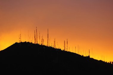 Canlı turuncu ve sarı günbatımı gökyüzüne karşı siluetlenmiş, dağların tepesindeki iletişim anten dizisi ve iletim kulelerinin çarpıcı silueti. Görüntü, bağlantı, teknoloji, altyapı ve doğa tarağının güzelliğini çağrıştırıyor.