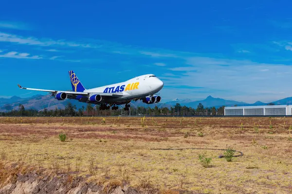Bir Atlas Air Boeing 747 kargo uçağı Quito 'nun Mariscal Sucre Havaalanı' ndaki piste inecek. Arka planda, Illinizas Dağları 'nın zirveleri ve bir hangar bu lojistik operasyonu açık bir gökyüzü altında çerçeveler..