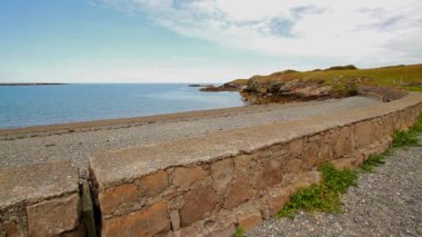 Anglesey, Galler 'in kuzey kıyısında Cemlyn Körfezi. Duvar, çakıl taşı plaj ve deniz, kayalık sahilli mavi gökyüzü.