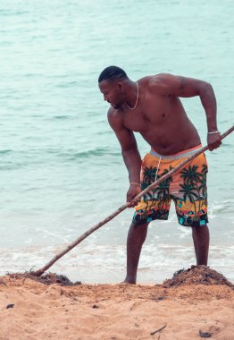 Samana, Dominican Republic, 26 august, 2022. Young and fit Dominican man cleaning the beach with a rake in beautiful and secluded Playa Ermitano.