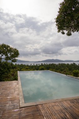 Denize ve diğer egzotik takımadalara bakan tropikal bir adadaki güzel sonsuzluk havuzunun geniş açılı görüntüsü. Bulutlu gün, neredeyse gün batımı, su yansımaları.