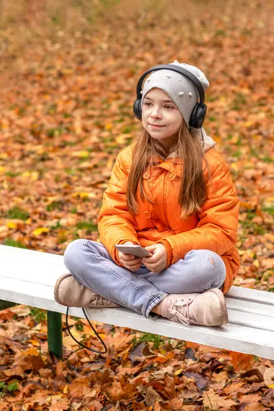 Vertical shot of a girl listening to music on headphones, sitting on a bench in an autumn park.