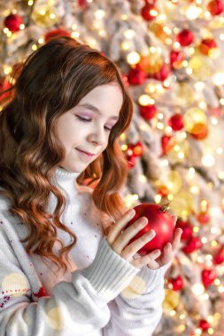 Bir kız süslenmiş bir Noel ağacının arka planında kırmızı bir yılbaşı balosuna bakıyor.