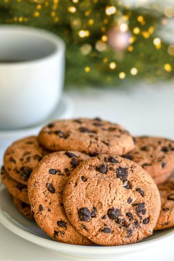 Bir tabakta çikolatalı kurabiyeler ve bir Noel ağacının arka planında bir fincan. Mutlu Noeller.
