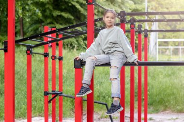 Bir fitness kızı güneş ışınlarında bir spor salonunun yatay çubuğunda oturur. Açık hava fitness.