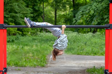 Bir fitness kızı, bir spor salonunun yatay çubuğunda baş aşağı asılı duruyor. Açık hava fitness.