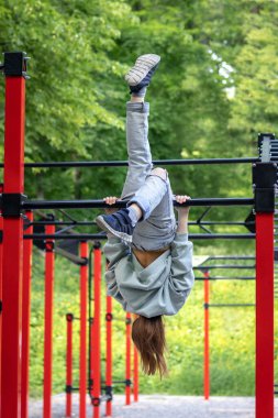 Bir fitness kızı, bir spor salonunun yatay çubuğunda baş aşağı asılı duruyor. Açık hava fitness.