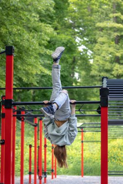 Bir fitness kızı, bir spor salonunun yatay çubuğunda baş aşağı asılı duruyor. Açık hava fitness.