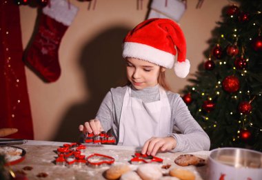 Noel Baba şapkalı bir kız Noel kurabiyelerini kesmek için kesicilerle çalışıyor.