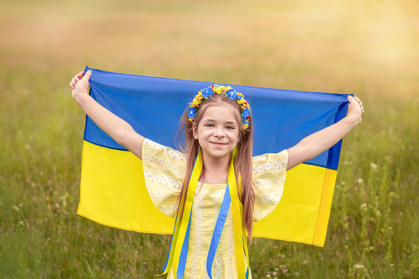 A girl in a wreath of yellow and blue flowers holds the flag of Ukraine in her hands against the background of a field. Ukrainian Independence Flag Day.