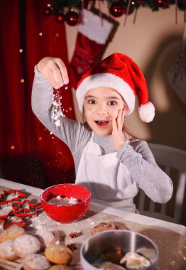 Noel Baba şapkalı bir kız şaşırtıcı bir şekilde Noel ağacının arka planına karşı masayı un ile kaplıyor.