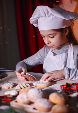 Şefin şapkasıyla neşeli kız Noel arkaplanında ev yapımı Noel kurabiyeleri yiyor.