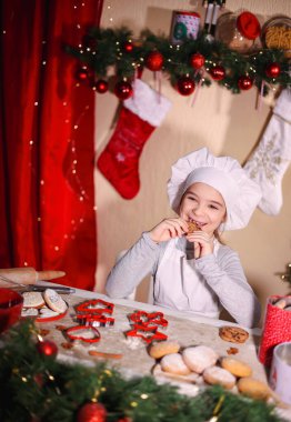 Şefin şapkasıyla neşeli kız Noel arkaplanında ev yapımı Noel kurabiyeleri yiyor.
