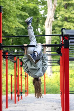 Genç bir insan yeşil doğayla çevrili renkli egzersiz çubuklarında baş aşağı sallanıyor..