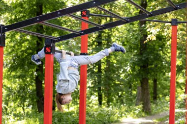 Bir çocuk yemyeşil bir parkta oyun parkında baş aşağı asılı duruyor..