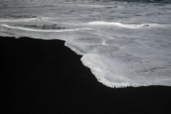 Mostly blurred black sand beach with white foam of sea waves. White and black background with copy space. Exotic black beach photo. Dark volcanic sand and white ocean waves