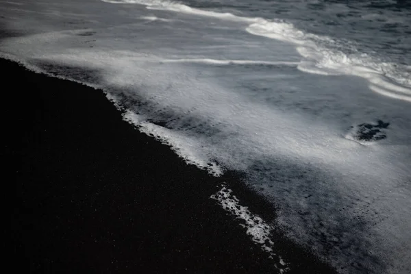 Mostly blurred black sand beach with white foam of sea waves. White and black background with copy space. Exotic black beach photo. Dark volcanic sand and white ocean waves