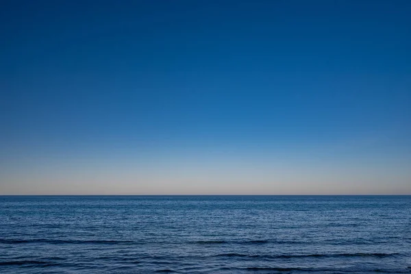 Blue ocean or sea background with a lot of blank space. Blue sky above calm water. Light waves in evening or sunset light. Minimalistic landscape. Marine view. Summer nature wallpaper. Vacation photo