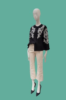 Full length image of a female display mannequin wearing black long sleeve blouse with embroidered pattern and white jeans isolated on a green background.