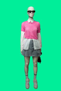 Yeşil arka planda pembe örgü bluz ve gri etek giyen bir kadın vitrin mankeni.