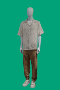 Yeşil arka planda kısa kollu gömlek ve kahverengi pantolon giyen bir kadın vitrin mankeninin tam boy görüntüsü