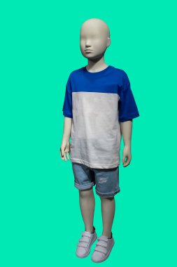 Yeşil arkaplanda iki renkli mavi-beyaz pamuklu tişört ve kot şort giyen bir çocuk mankeninin tam boy görüntüsü