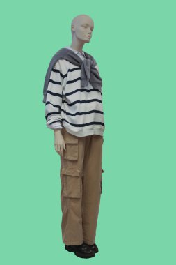 Yeşil arka planda beyaz çizgili kazak ve kahverengi kargo pantolon giyen bir kadın vitrin mankeni.