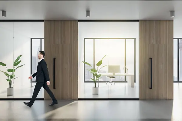 Businessman walking in modern office hallway interior with wooden, concrete and glass elements, window with city view and daylight. decorative plants