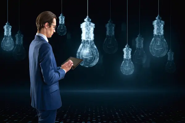 Attractive young european businessman using smartphone with glowing digital lamp hologram on dark background. Idea, innovation and future concept