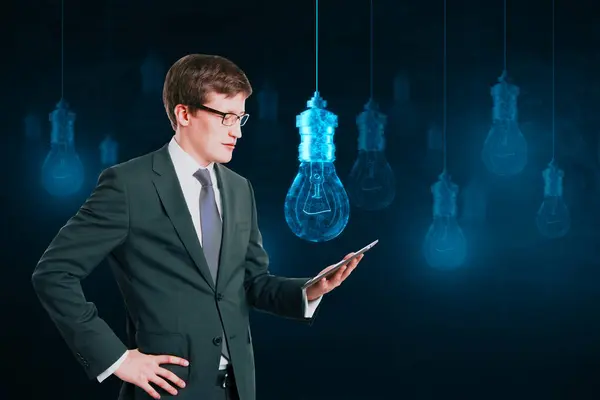 Attractive young european businessman using cellphone with glowing digital lamp hologram on dark background. Idea, innovation and future concept