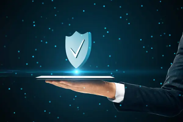 Internet privacy and cyber protection concept with check mark on blue shield above digital tablet in man hand on abstract dark background with dots