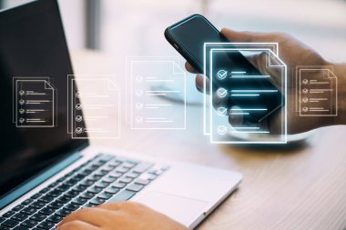 Ofis arka planında yaratıcı dijital belge bulunan cep telefonu ve dizüstü bilgisayarı kullanarak iş adamının elini kapat. Çevrimiçi inceleme ve dijital form kontrol listesi. Oylama konsepti. Çift pozlama.