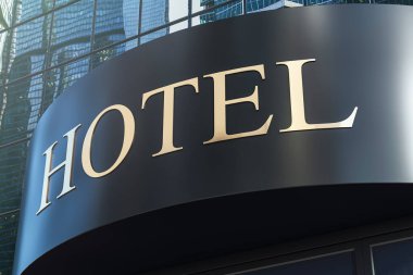 Arka planda cam gökdelen yansımaları olan parlak, siyah bir dış cephede altın harflerle bir otel tabelasının yakın görüntüsü. 3B Hazırlama