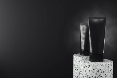 Terrazzo podyumlarında iki siyah kozmetik tüp, gerçekçi 3D görüntüleme, koyu dumanlı arka plan, ürün markalaştırma, reklam ve sunum konsepti