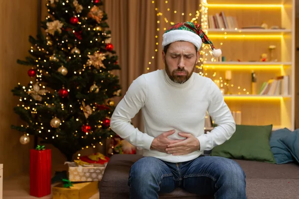 The man is sick at home, has a severe stomach ache, sits on the couch near the Christmas tree, is alone at home celebrating the New Year in a New Years hat, holding his stomach with his hands.
