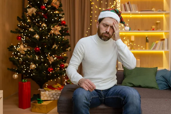 Sad man on Christmas alone for two, sitting on the sofa at home in depression near the Christmas tree after celebrating the new year.