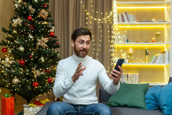 Businessman at home for Christmas and New Year holidays sitting on sofa in living room near Christmas tree talking on video call, man having fun talking with friends holding phone