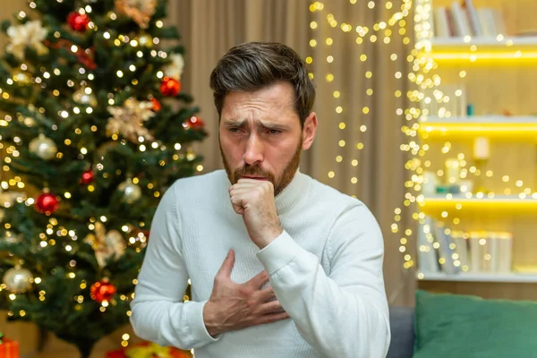 man coughs on christmas at home sitting alone on sofa sick on new year holidays in living room.