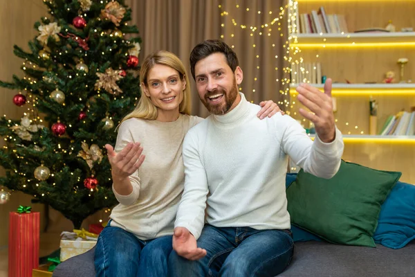 Video call family couple, a man and a woman, are talking to friends, remotely looking at the web camera, and waving and greeting, inviting friends to visit to celebrate Christmas and the New Year.