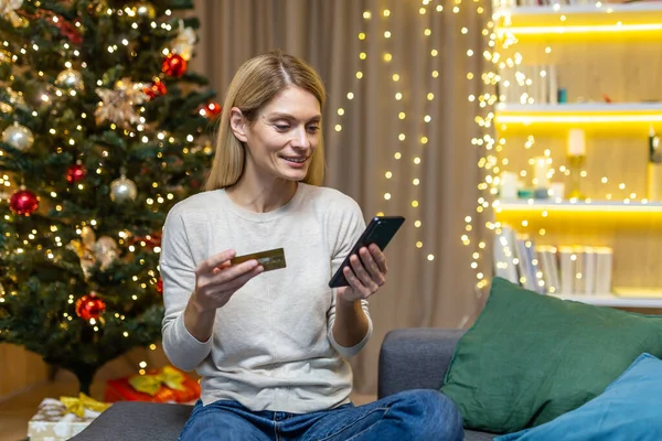 Happy woman alone at home buying Christmas gifts online, housewife sitting on sofa near Christmas tree holding phone and bank credit card, remote shopping for new year.