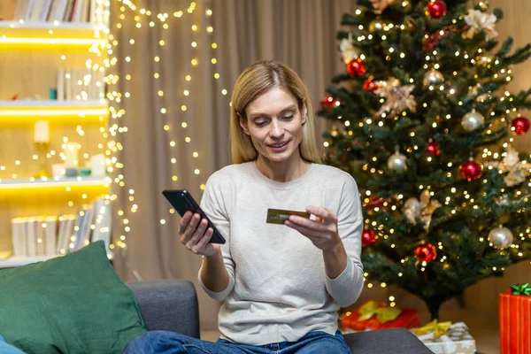 Happy woman alone at home buying Christmas gifts online, housewife sitting on sofa near Christmas tree holding phone and bank credit card, remote shopping for new year.