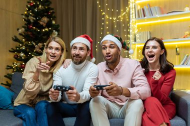 Noel tatilinde eğlenen ve kutlayan dört farklı arkadaşından oluşan yeni yıl partisi grubu, koltukta oturan erkekler ve kadınlar oyun konsollarında oyun oynuyorlar..